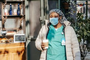 Adult black woman in nurse uniform with jacket and protective mask with headwear in city street with cup of drink near counte