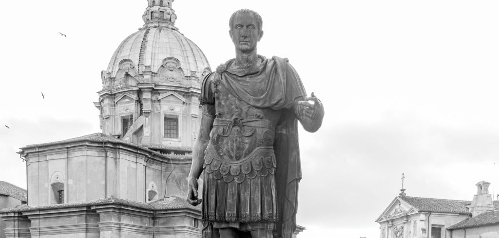 Black and white photo of Julius Caesar's statue with Roman architecture in the backdrop. — Julius Caesar The Man Who Changed