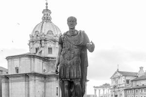 Black and white photo of Julius Caesar's statue with Roman architecture in the backdrop. — Julius Caesar The Man Who Changed